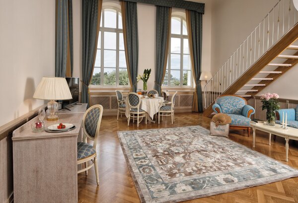 Ein elegantes Hotelzimmer mit großen Fenstern, die viel Tageslicht hereinlassen. Im Raum befinden sich ein Esstisch mit Stühlen, ein gemütlicher Sitzbereich mit blauen Sofas und ein Teppich auf dem... Kaiserliche Suiten im Hotel mit Meerblick Kroatien