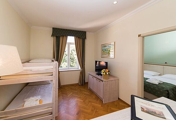 Ein helles Hotelzimmer mit einem Etagenbett und einem Doppelbett. Ein Fenster mit Vorhängen lässt Tageslicht herein. Ein Fernseher und eine Blumenvase stehen auf einem Holzschrank. Der Raum hat ein... Wunderschöne Zimmer im Familienhotel Crikvenica